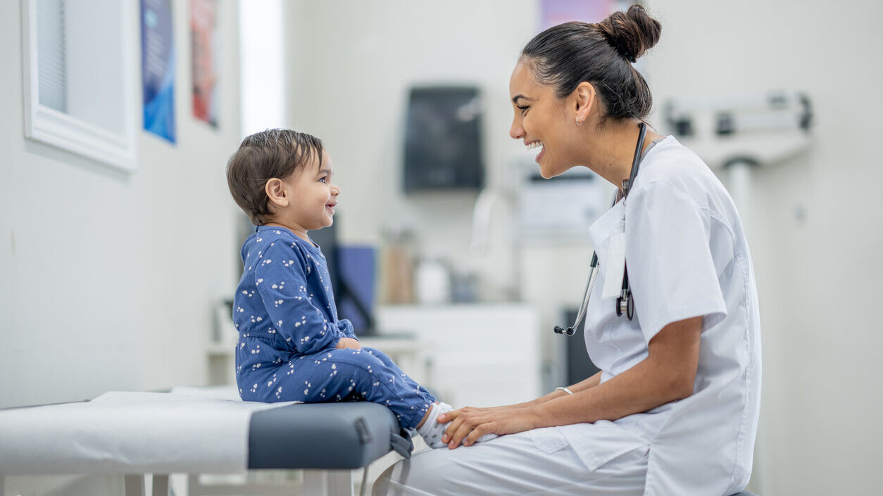 Decorative image. A doctor in hospital clothes is squatting in front of a little boy in a blue romper. They are smiling at each other.