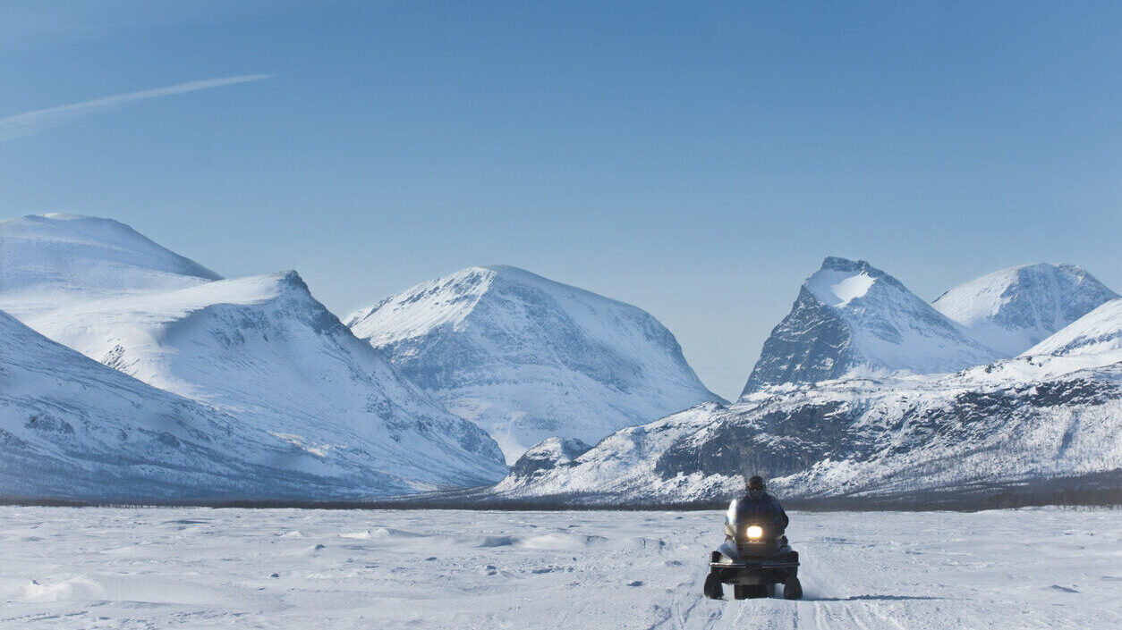 Dekorativ bild. Snötäckt landskap med Kalix berg och en klarblå himmel i bakgrunden. I förgrunden syns en ensam man på snöskoter.